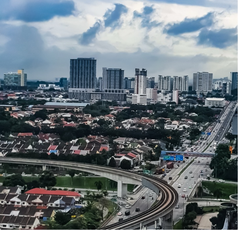 Aerial view of a modern city with high-rise apartment and office buildings in the background, a residential neighborhood with red-roof houses in the foreground, and an elevated rail line curving alongside a busy multi-lane highway under cloudy skies.